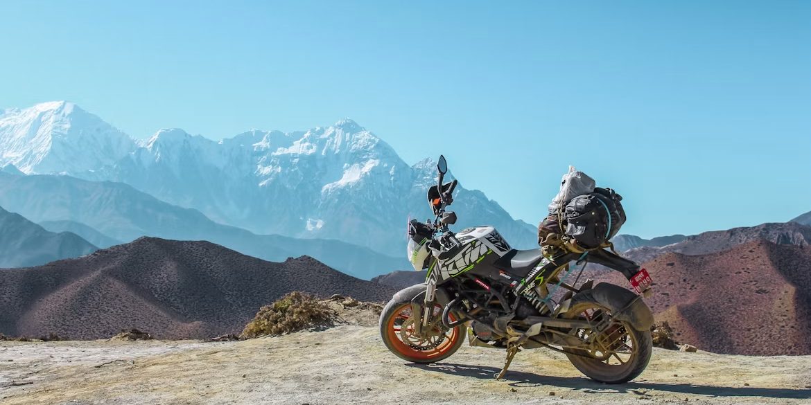 An adventure motorcycle standing on a bare rock ridge above arid mountain country, with a snow topped range on the far horizon