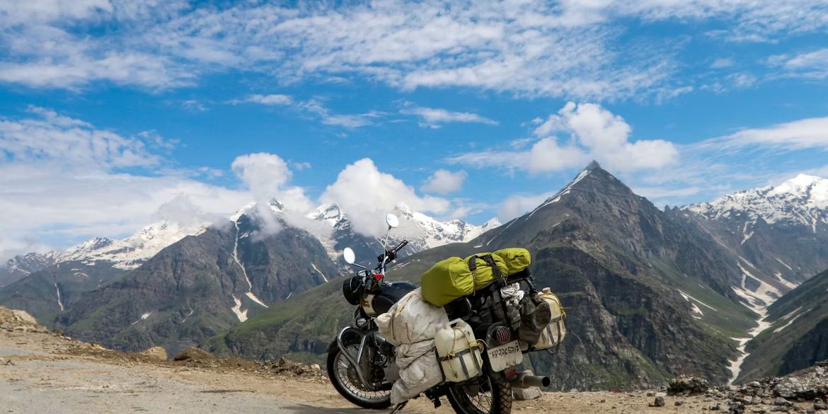A loaded adventure motorcycle with a dry bag strapped over the rear, parked on gravel high ground with snow topped peaks behind