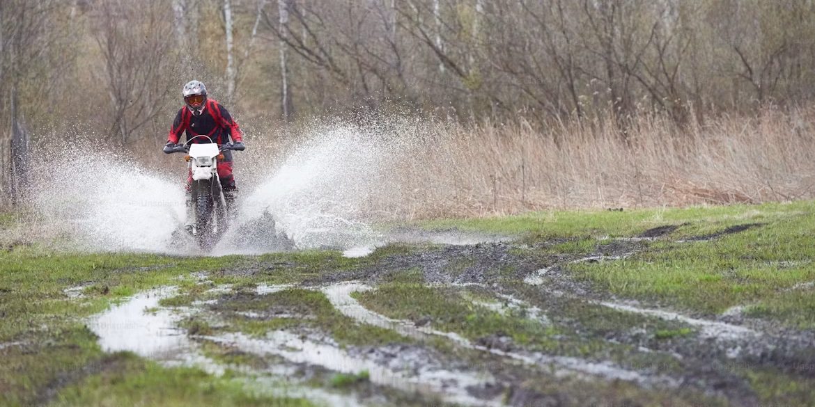 A rider crossing a flooded dirt track and throwing a wide sheet of spray, with scrub and bare trees on both sides