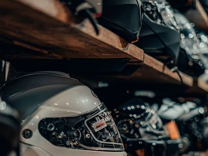 A road helmet resting beneath a heavy timber shelf in a dim workshop, with a second helmet stored on the shelf above