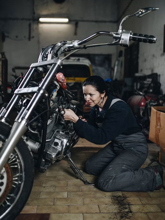 A mechanic in dark overalls kneeling beside a motorcycle in a workshop bay, working at the engine and exhaust