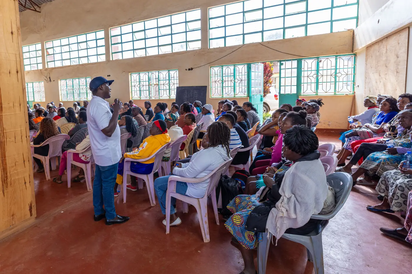 A man in a HerNext Network shirt speaking to a hall full of women seated on plastic chairs