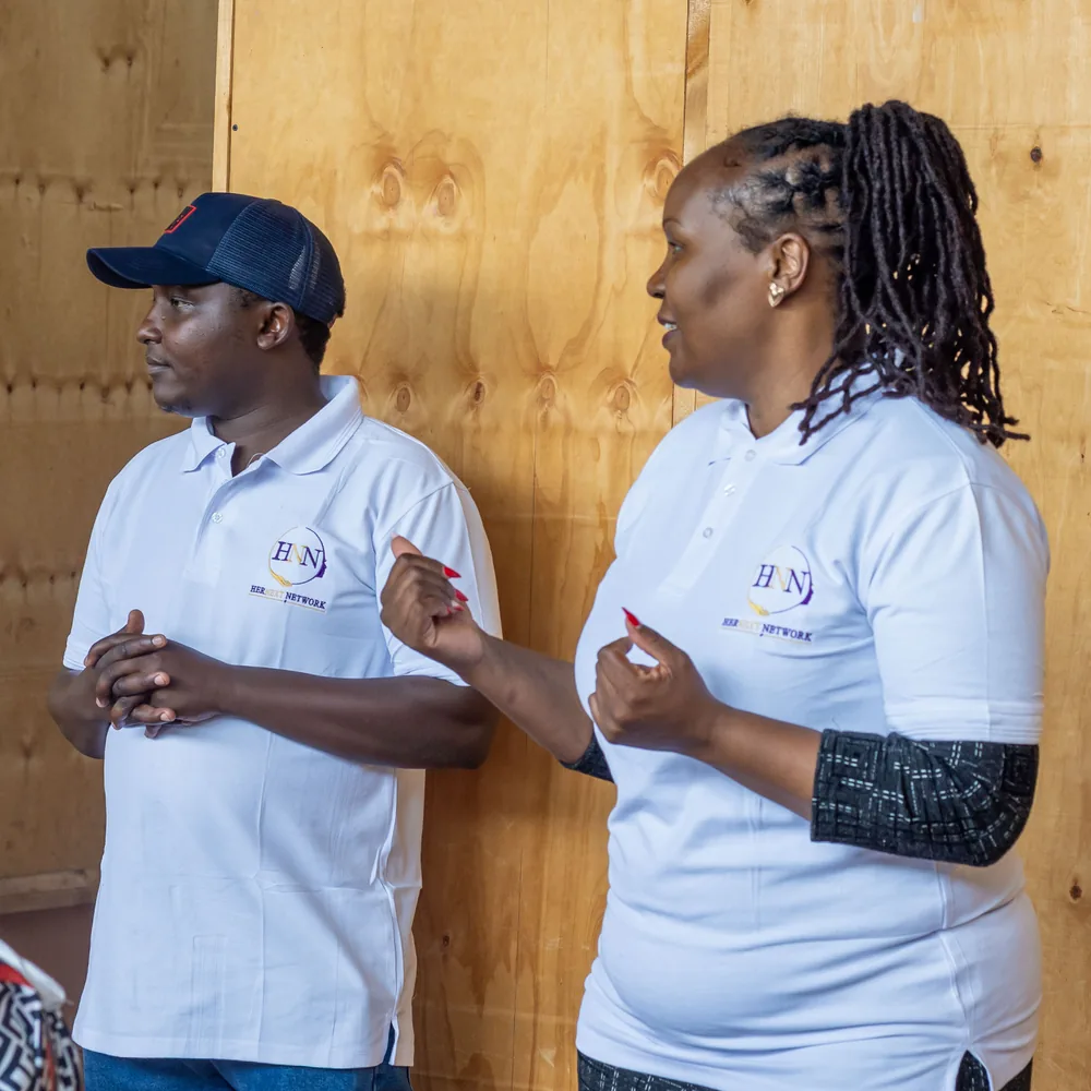 Two people in HerNext Network shirts turned towards the room, the woman gesturing as she speaks