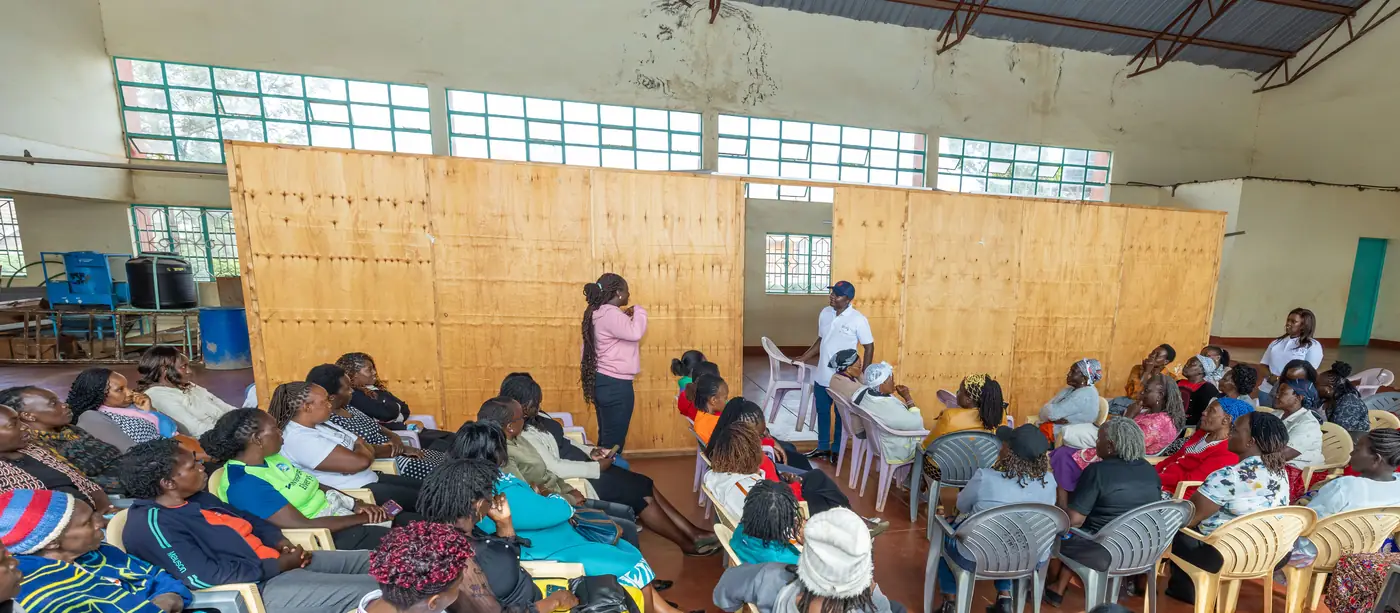 A wide view of a community hall, rows of women seated facing two people standing at a plywood partition