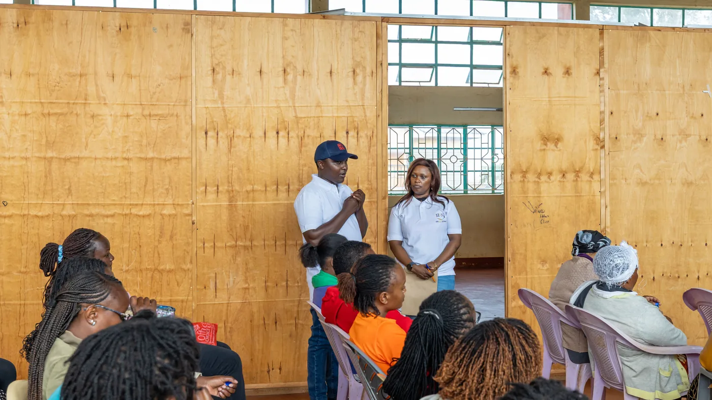 Two people in HerNext Network shirts standing to address a seated group in a community hall