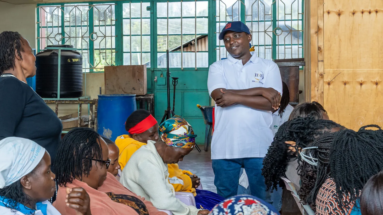 A man in a HerNext Network shirt, arms folded, smiles at a woman standing among seated women in a community hall