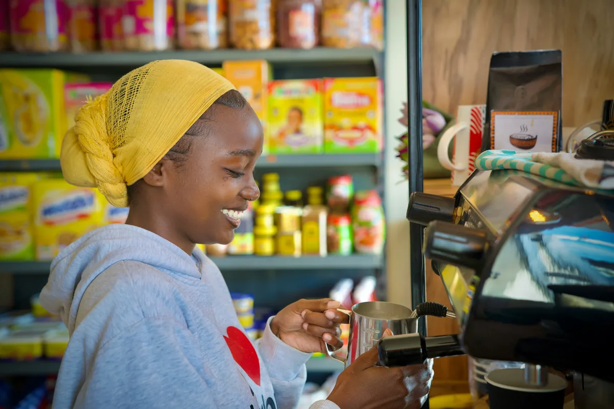A smiling young woman in a yellow headwrap preparing coffee at an espresso machine in a small shop
