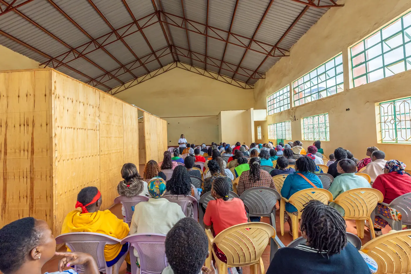 Rows of women seated in a community hall, seen from the back, facing a speaker at the far end